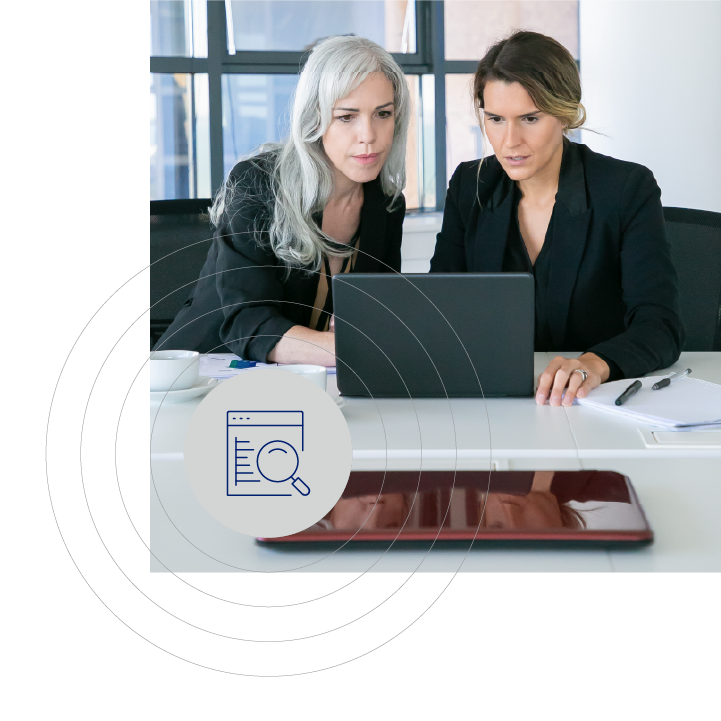 Two women in business attire seated at a white table, focused on a laptop, with a web browser graphic overlay. The scene includes documents and writing instruments.